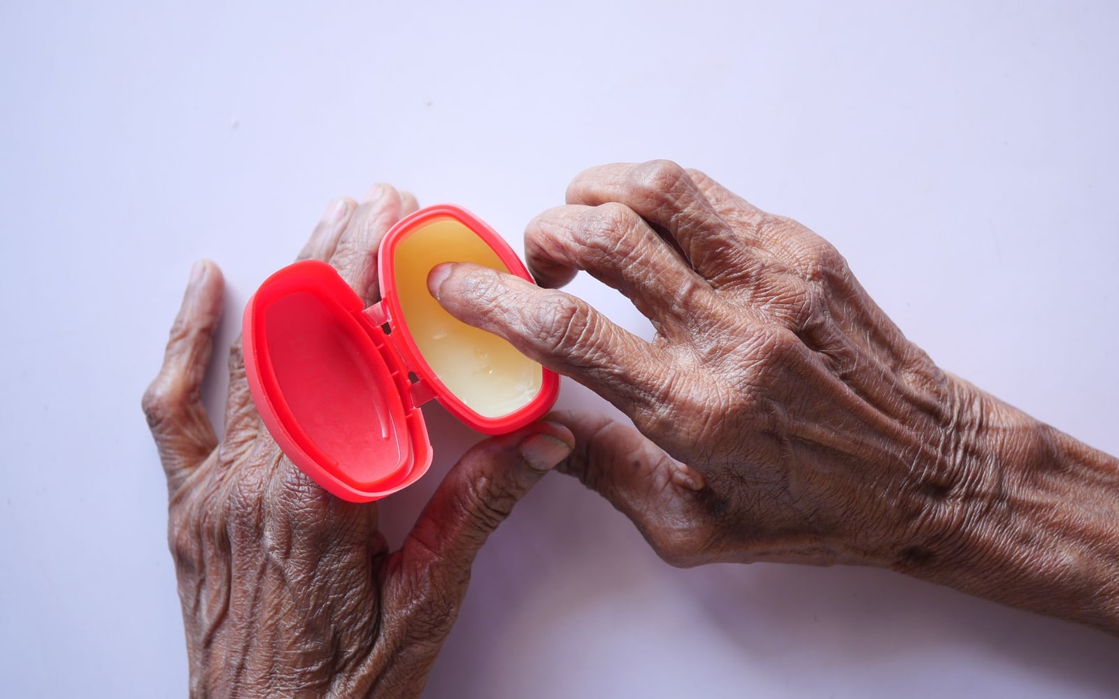 Close-up of senior hands using hand cream for skincare in a red container on a white background.
